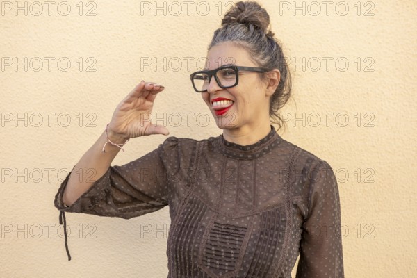 A woman with glasses and red lipstick enthusiastically demonstrates a sign language gesture outdoors against a soft yellow wall She exudes positivity and confidence in communication