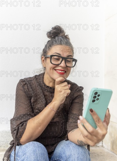 A woman with glasses uses a smartphone for a video call, focusing on learning sign language She is seated against a light background, exuding enthusiasm and engagement