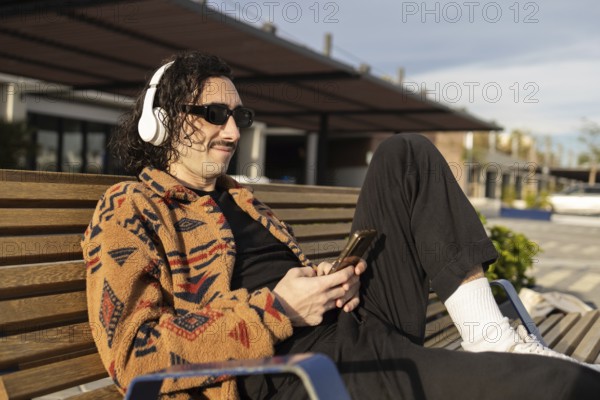 A man relaxes on a bench, wearing headphones and sunglasses, enjoying music on a sunny day He wears a patterned fleece jacket, black pants, and holds a smartphone