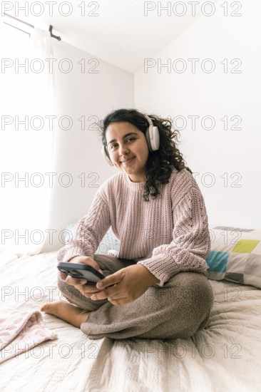 A young latin woman enjoying leisure time at home, wearing headphones, and using a smartphone while sitting on a comfortable bed Bright, casual interior setting