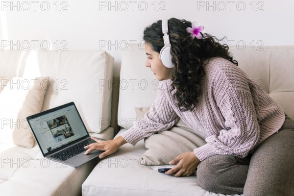 A young latin woman relaxes on a cozy sofa at home, wearing headphones and browsing her laptop She holds a phone, enjoying her leisure time in a comfortable sweater