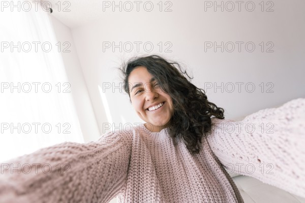 A cheerful young latin woman with curly hair wears a cozy pink sweater, smiling as she enjoys a peaceful day at home with natural light streaming in through the window