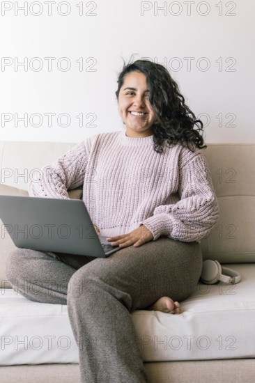 A cheerful young hispanic woman sits comfortably on a sofa at home, using her laptop She is dressed casually, appearing relaxed and content in the cozy environment