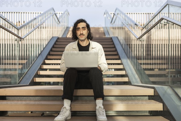 A man sits on an outdoor stairway, focused on his laptop Dressed casually, he embodies a modern, tech-savvy lifestyle, balancing work and relaxation in an urban setting