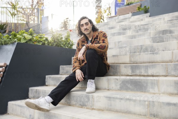 A man sits on outdoor steps, showcasing a relaxed, casual urban style Wearing a patterned jacket, black pants, and sneakers, he exudes a modern, effortless fashion vibe