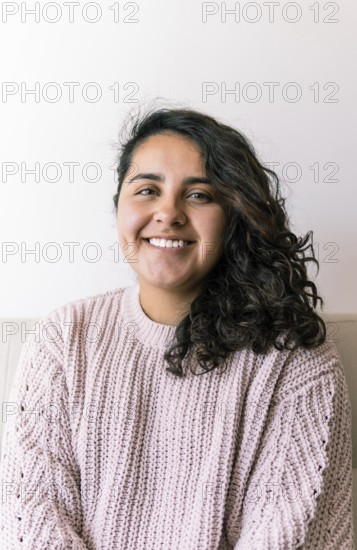 A young latin woman with curly hair smiles warmly, sitting indoors wearing a cozy sweater The relaxed atmosphere captures a candid moment of comfort and happiness at home