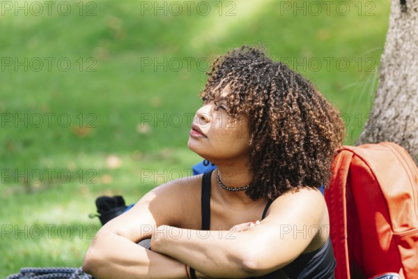 A Black woman sits under a tree in a Bogota park, resting her arms on her knees while gazing into the distance Surrounded by greenery, she appears reflective and calm