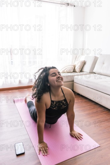 A young latin woman performs yoga on a pink mat in her cozy living room, enjoying a peaceful moment Sunlight streams through curtains, creating a relaxed ambiance