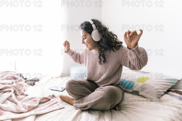 A young latin woman sits cross-legged on her bed, wearing headphones and a cozy sweater She appears relaxed and happy, enjoying music in a bright, comfortable room