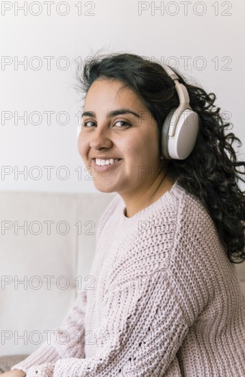 A young latin woman sits on a cozy couch, wearing headphones and a knitted sweater She is smiling, exuding a sense of relaxation and joy, capturing a moment of peaceful leisure