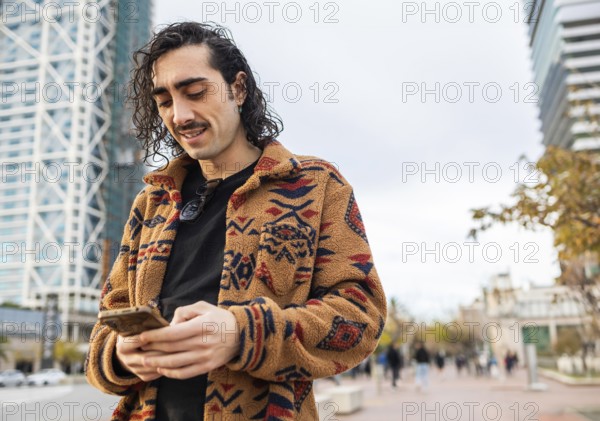 A fashionable man with curly hair wearing a patterned jacket checks his smartphone on a city street Modern buildings are in the background, showcasing a vibrant urban lifestyle