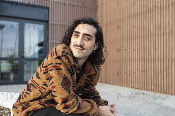A man with long hair and a mustache, wearing a cozy patterned jacket, smiles while sitting outdoors The background shows a modern building facade, adding urban charm