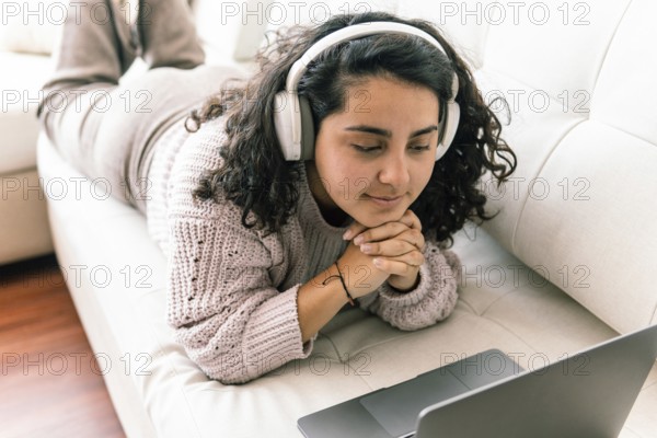 A young latin woman relaxes on a couch, wearing headphones and a cozy sweater, as she engages with her laptop The setting exudes comfort and tranquility in a home environment