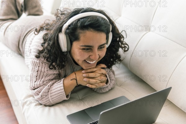 A young latin woman wearing headphones lies on a sofa, smiling warmly as she watches something on her laptop Cozy sweater and casual home setting reflect relaxation and contentment