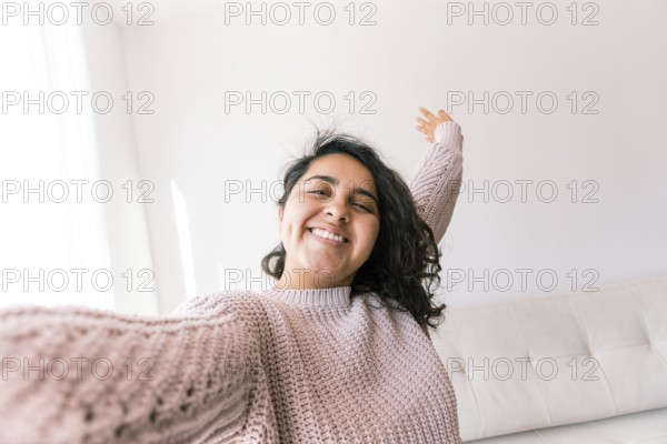 A young latin woman smiles joyfully while stretching in her comfortable home environment She wears a cozy sweater and enjoys the warm natural light coming through the window