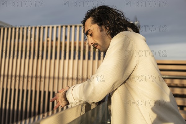 A man in a light shirt leans on a railing, captured in soft evening light His thoughtful expression and the wooden background create a serene and reflective atmosphere
