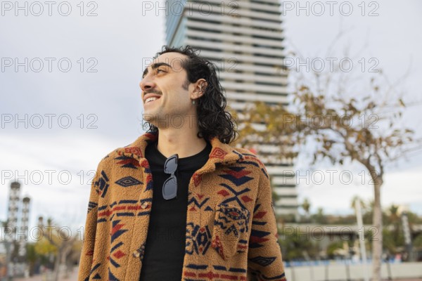 A stylish man with a patterned coat and sunglasses smiles confidently in an urban setting A modern building and trees provide an appealing backdrop