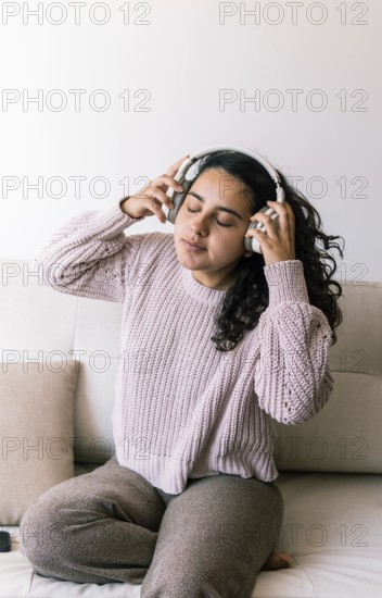 A young latin woman with curly hair relaxes on a couch at home, wearing a cozy sweater and headphones She enjoys listening to music with her eyes closed