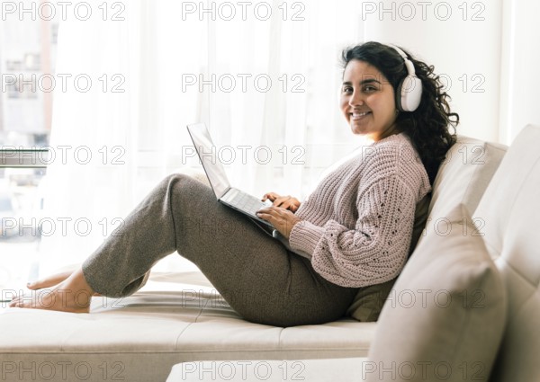 A young latin woman in cozy attire sits comfortably on a sofa at home, using a laptop with headphones on She exudes relaxation and focus, enjoying a peaceful indoor environment