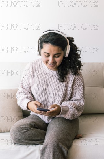 A young latin woman sits comfortably on a couch, wearing headphones and looking at her phone She exudes relaxation and happiness, enjoying her time at home