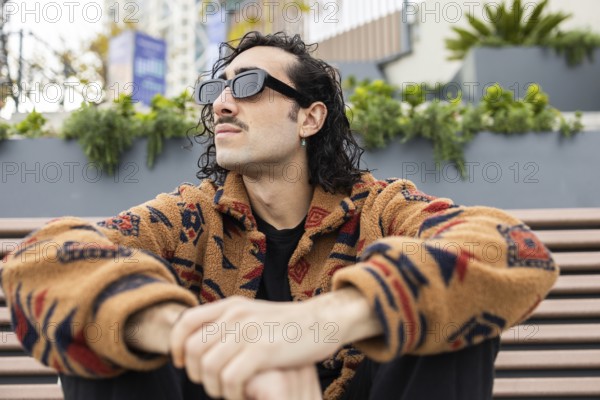 A fashionable man wearing sunglasses and a patterned jacket sits on a bench outdoors, exuding a relaxed and confident vibe in an urban environment
