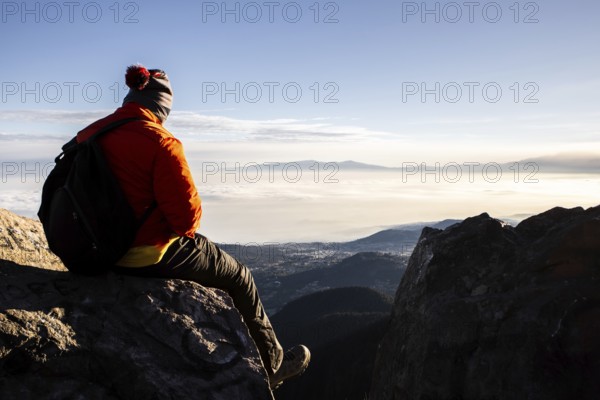 Back view of unrecognizable man sitting atop a mountain during a breathtaking sunrise, equipped with a hiking backpack, against a background of vast clouds and distant peaks