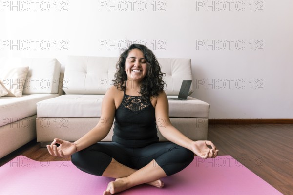 A young latin woman sits peacefully on a yoga mat in a serene living room She practices yoga, focusing on relaxation and mindfulness, embracing a moment of calm and tranquility