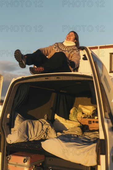 A Latina woman laughs joyfully as she sits in the open trunk of a car, surrounded by luggage and blankets, during a sunset in Iceland. She is dressed warmly and appears relaxed and happy
