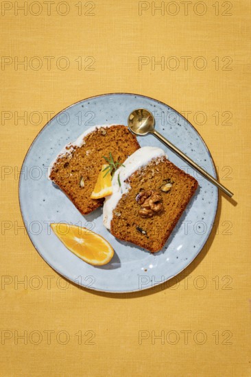 Slices of carrot cake loaf topped with cream cheese frosting, garnished with walnuts, rosemary, and orange wedges on a blue plate, set on a yellow linen background