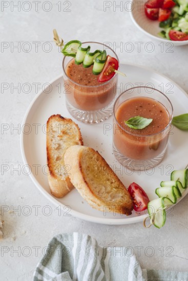 Two glasses of refreshing gazpacho are garnished with cucumber and tomato skewers, accompanied by slices of crusty bread, served on a light, textured table setting
