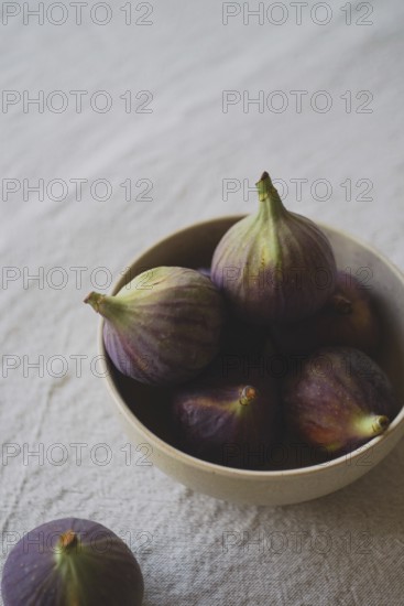 A rustic ceramic bowl filled with ripe, purple figs placed on a textured linen tablecloth, capturing a natural and earthy ambiance, ideal for culinary-themed designs