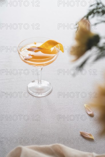 A sophisticated mandarin cocktail adorned with a vibrant orange peel zest, served in a classic stem glass on a textured white background, accompanied by soft natural light and subtle floral accents