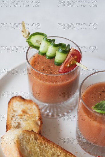 A refreshing glass of gazpacho garnished with cucumber, tomato, and bell pepper on a skewer Served with slices of grilled bread, perfect for a summer appetizer