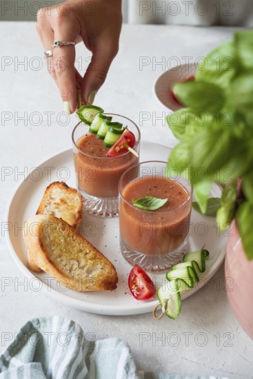 A hand garnishes gazpacho with cucumber and tomato skewers in two glass cups The plate also has toasted bread slices A fresh and appetizing presentation with herbs