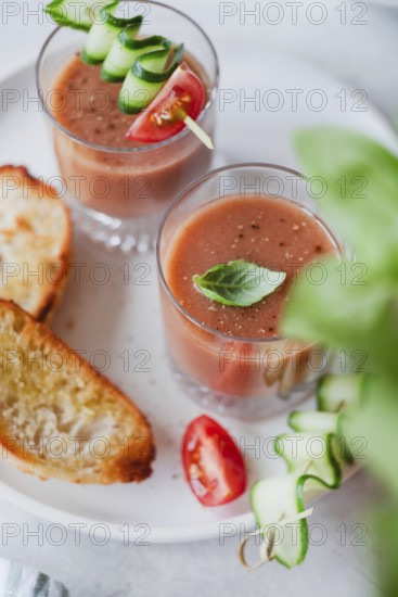 Two glasses of gazpacho topped with cucumber and tomato skewers, alongside toasted bread, served on a white plate Perfect for a light, refreshing appetizer or snack