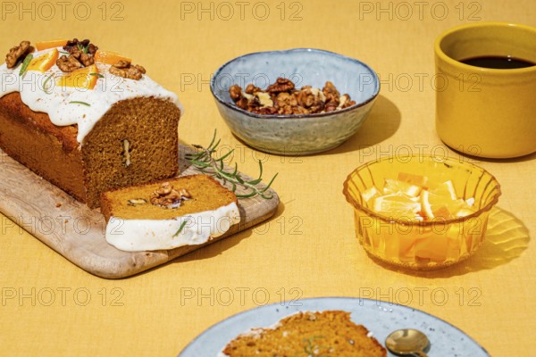 A moist carrot cake loaf topped with cream cheese frosting, garnished with walnuts and rosemary Served on a wooden board, with orange slices, coffee, and walnuts on a yellow table