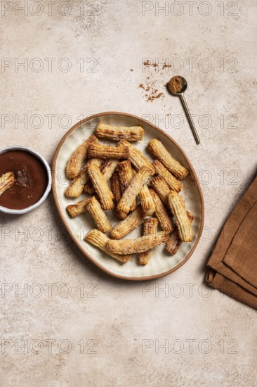 Top view of a ceramic plate filled with homemade churros, coated with sugar and cinnamon, accompanied by a bowl of chocolate sauce and a sprinkling of cinnamon, presented on a textured background