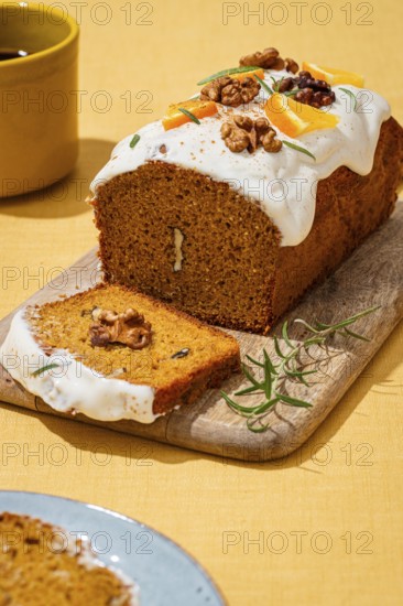 A freshly baked carrot cake loaf adorned with smooth cream cheese frosting, walnut pieces, and aromatic rosemary Served on a rustic wooden board over a linen cloth