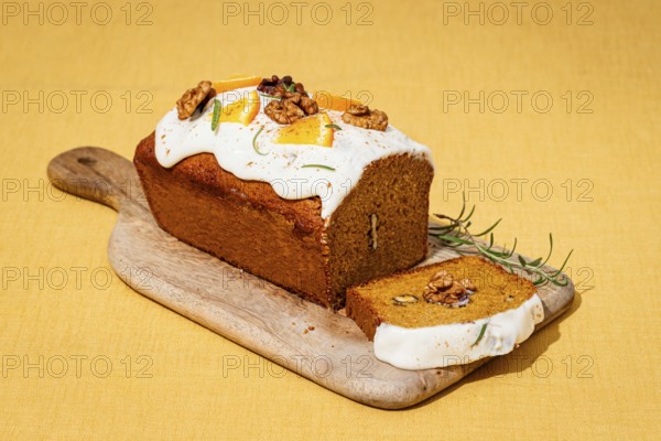 A freshly baked carrot cake loaf topped with cream cheese frosting, garnished with walnuts and rosemary Set on a wooden board against a yellow linen backdrop