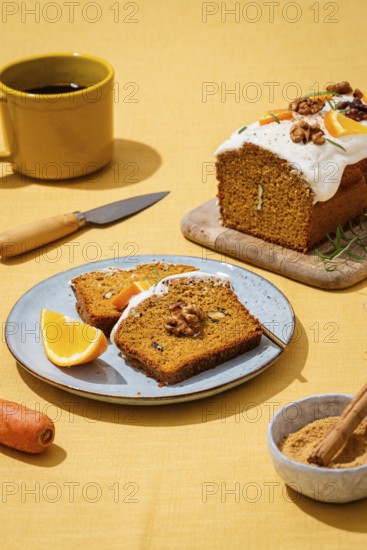 A tasty carrot cake loaf topped with creamy frosting and walnuts, presented on a yellow linen tablecloth Garnished with orange wedges and rosemary sprigs