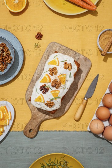 Carrot cake loaf topped with cream cheese frosting, orange slices, walnut, and rosemary Set on a wooden board with yellow linen background featuring baking essentials
