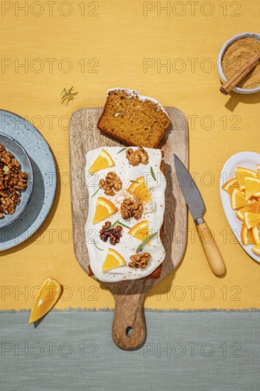 A delicious carrot cake loaf topped with creamy cream cheese frosting, garnished with orange slices, walnuts, and rosemary on a wooden board with a yellow linen backdrop