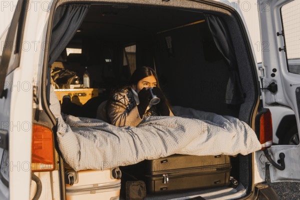 A Latina female relaxes with a coffee in a cozy van setup during a picturesque sunrise at Camp Iceland. She is looking away from the camera, immersed in the tranquil moment