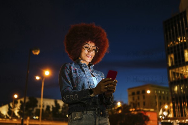 African descendent woman with afro hair, wearing glasses and a denim jacket, uses her smartphone on a city street at night, illuminated by streetlights
