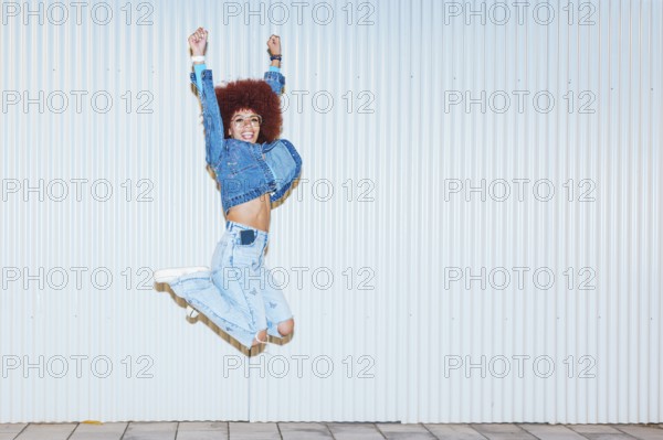 A joyful African descent woman with afro hair and fashionable denim outfit jumping energetically against a corrugated white background