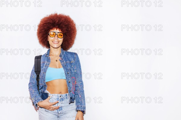 African descendant woman with a vibrant afro hair, smiling while wearing orange sunglasses and a denim jacket, standing against a plain white background
