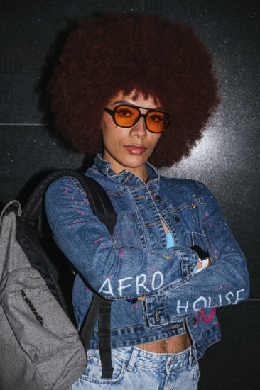 African descent woman with a voluminous afro hairstyle wearing orange sunglasses and a denim jacket, confidently posing against a dark background with arms folded and looking at camera