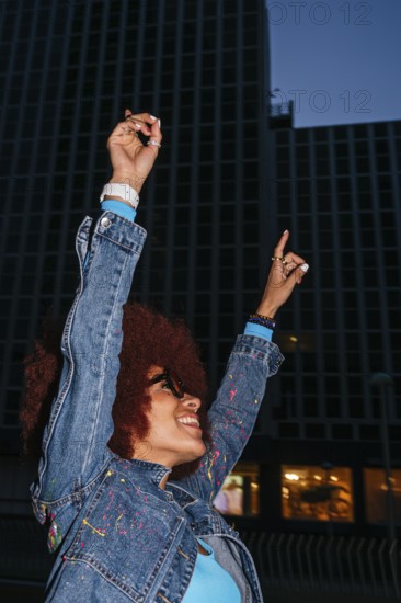 A joyful African descendant woman with afro hair raises her arms in celebration against a backdrop of an urban skyscraper at night. She is wearing a denim jacket and is captured smiling broadly, making a victory gesture