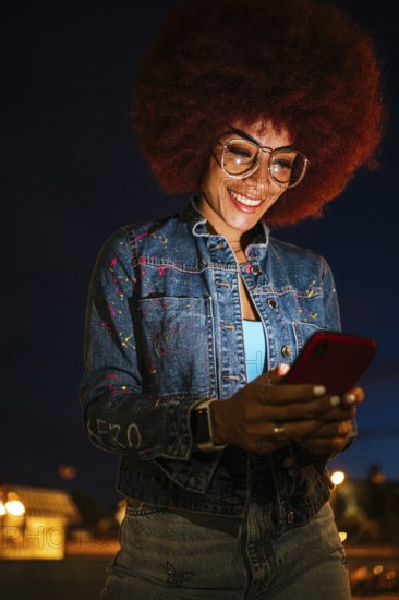 African descendent woman with a vibrant afro hairstyle, wearing glasses, joyfully engaging with her smartphone. Her fashionable denim attire and brightly lit backdrop highlight her presence during a vibrant night scene