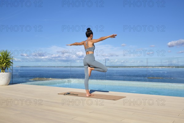 A woman practices yoga on a deck overlooking the ocean. She is in a balance pose, embracing tranquility under a clear blue sky by a poolside